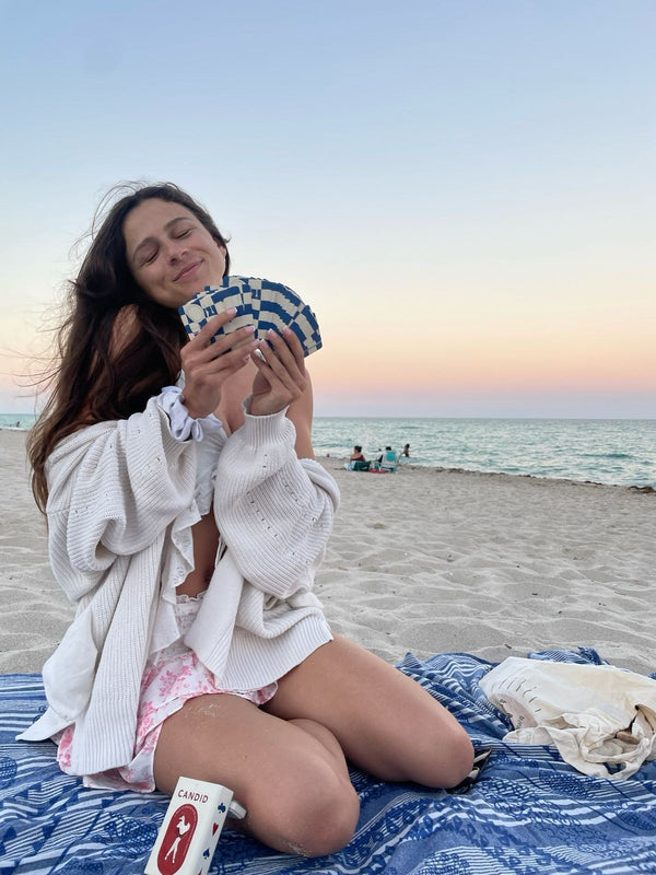 A woman on a checkered blanket at the beach at sunset holding CANDID cards